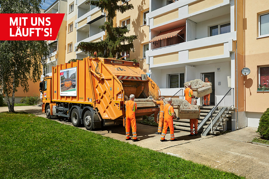 Zwei Mitarbeiter der HWS in orangener Kleidung tragen ein altes Sofa aus einem Hauseingang eines Plattenbaus.